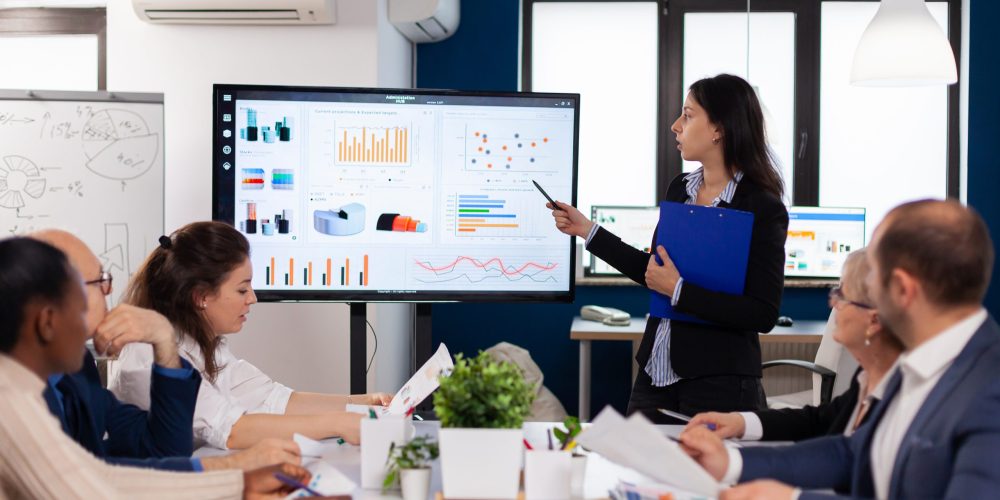 Young team leader in big corporation briefing coworkers pointing at graph meeting. Corporate staff discussing new business application with colleagues looking at screen